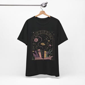 T-shirt featuring books, flowers, a snake, and stars framed by a mystical arched window—celestial, witchy, and dark academia styled.
