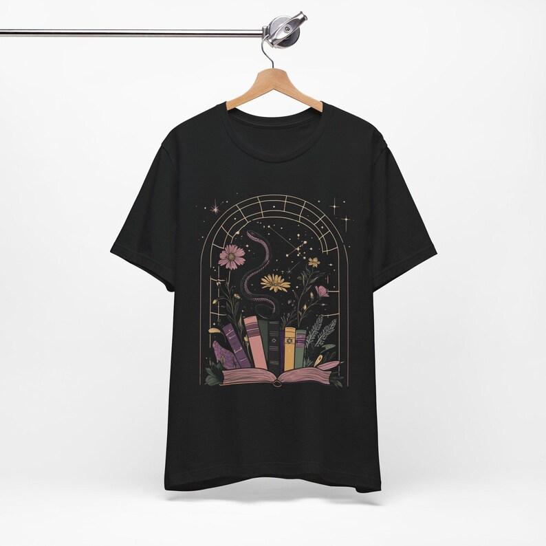 T-shirt featuring books, flowers, a snake, and stars framed by a mystical arched window—celestial, witchy, and dark academia styled.