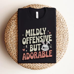 Funny graphic T-shirt featuring a sassy bunny with the phrase "Mildly Offensive But Adorable," perfect for humor-loving animal lovers. The main colors are white, beige and peach.