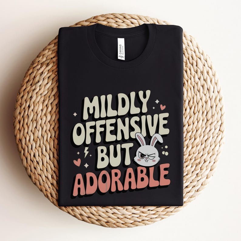 Funny graphic T-shirt featuring a sassy bunny with the phrase "Mildly Offensive But Adorable," perfect for humor-loving animal lovers. The main colors are white, beige and peach.
