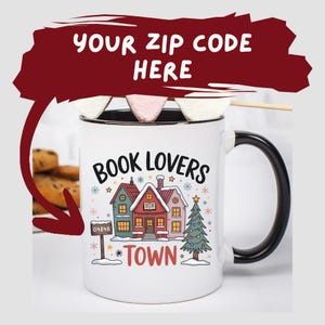 May include: A white ceramic mug with a black handle and rim, featuring the text "BOOK LOVERS TOWN" and a winter village scene. The mug has a marshmallow and a wooden stick on top.