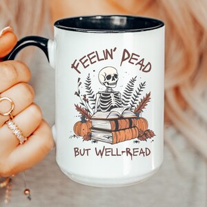 A white mug with a black or pink handle featuring a skeleton reading a book, surrounded by fall leaves and pumpkins, with the text: Feelin' Dead But Well-Read. Main colors are brown and orange.