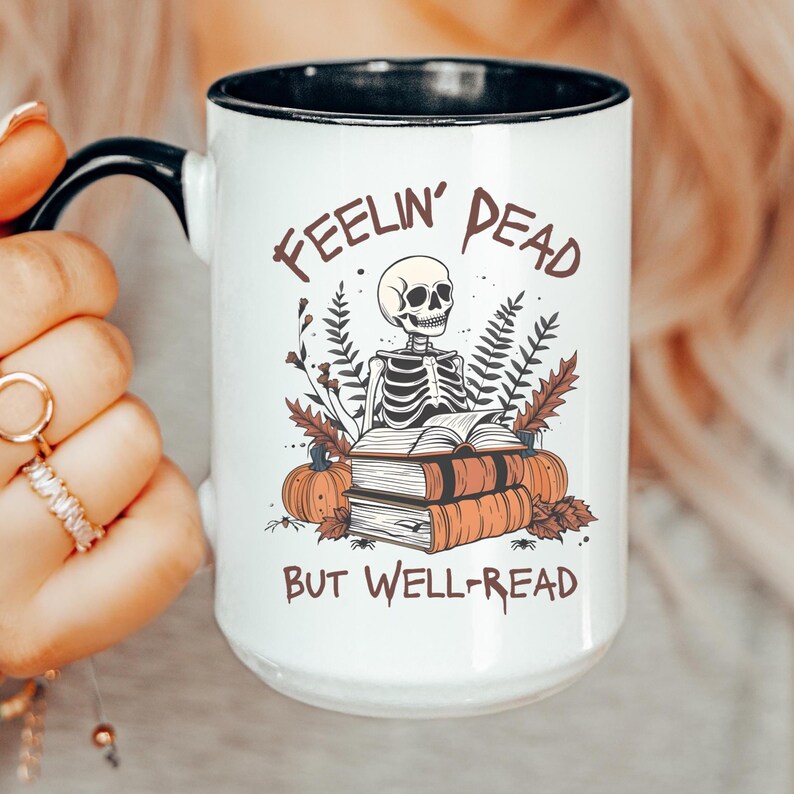 A white mug with a black or pink handle featuring a skeleton reading a book, surrounded by fall leaves and pumpkins, with the text: Feelin' Dead But Well-Read. Main colors are brown and orange.