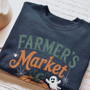 Könnte beinhalten: Marineblaues Sweatshirt mit dem Schriftzug "FARMER'S Market" in Grün und Orange. Das Design zeigt einen Kürbis, einen Geist, eine Spinne und Spinnweben. Das Sweatshirt liegt gefaltet auf einer Holzoberfläche.