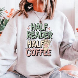 Könnte beinhalten: Ein hellgraues Sweatshirt mit dem Text "Half Reader Half Coffee" in bunten Buchstaben. Neben dem Text befindet sich eine Comic-Kaffeetasse mit einem Herzen.