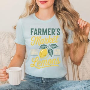 T-shirt with vintage-style “Farmer’s Market Lemons” graphic featuring a yellow lemon with leaves and rustic text. The main colors are yellow and green.