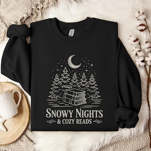 Puede incluir: Sudadera negra con un gráfico blanco de una luna creciente, estrellas, pinos y libros. El texto dice "Snowy Nights & Cozy Reads". La sudadera tiene mangas largas.