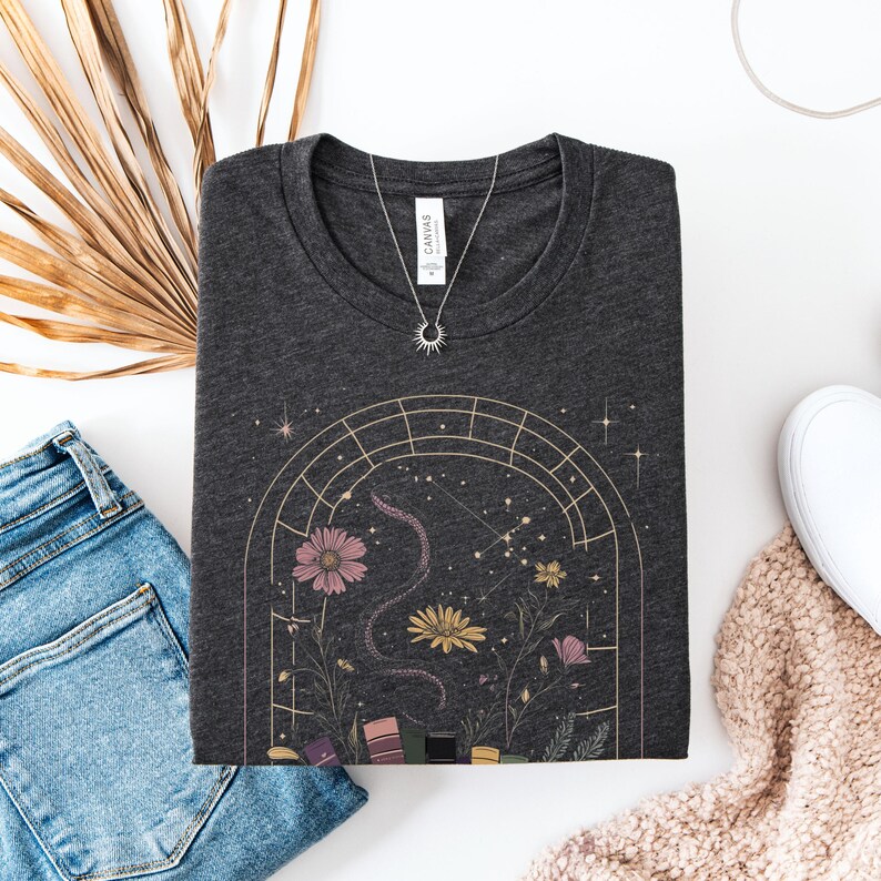T-shirt featuring books, flowers, a snake, and stars framed by a mystical arched window—celestial, witchy, and dark academia styled.