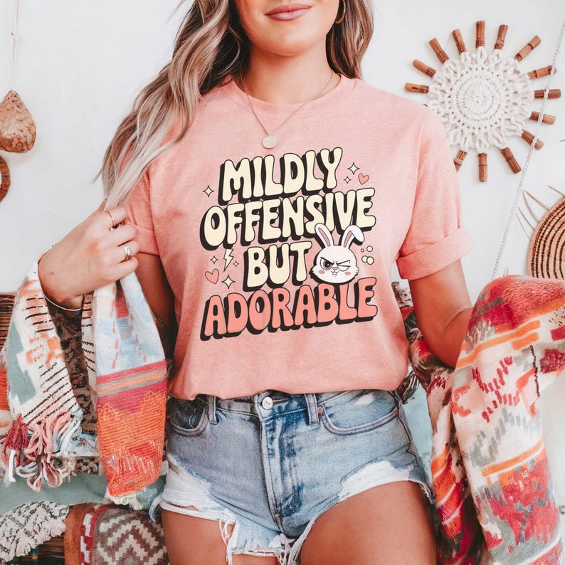 Funny graphic T-shirt featuring a sassy bunny with the phrase "Mildly Offensive But Adorable," perfect for humor-loving animal lovers. The main colors are white, beige and peach.