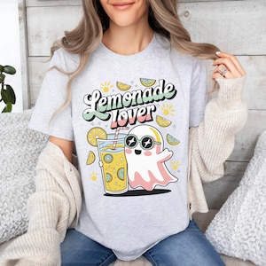 Peut inclure: T-shirt gris clair avec l'inscription "Lemonade Lover" dans une police r&eacute;tro. Le t-shirt pr&eacute;sente un fant&ocirc;me de dessin anim&eacute; portant des lunettes de soleil et tenant un verre de limonade. Des illustrations de citron et de soleil sont &eacute;galement pr&eacute;sentes.