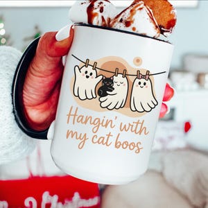 Könnte beinhalten: Weiße Keramiktasse mit schwarzem Rand, mit einem Design von drei Cartoon-Geistern und einer schwarzen Katze, die an einer Wäscheleine hängen. Der Text "Hangin' with my cat boos" ist auf die Tasse gedruckt. Die Tasse ist mit einem heißen Getränk gefüllt und mit Marshmallows belegt.