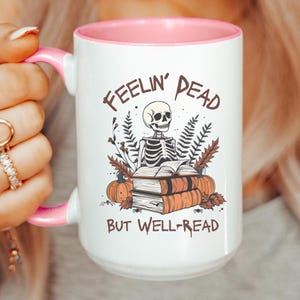 A white mug with a black or pink handle featuring a skeleton reading a book, surrounded by fall leaves and pumpkins, with the text: Feelin' Dead But Well-Read. Main colors are brown and orange.
