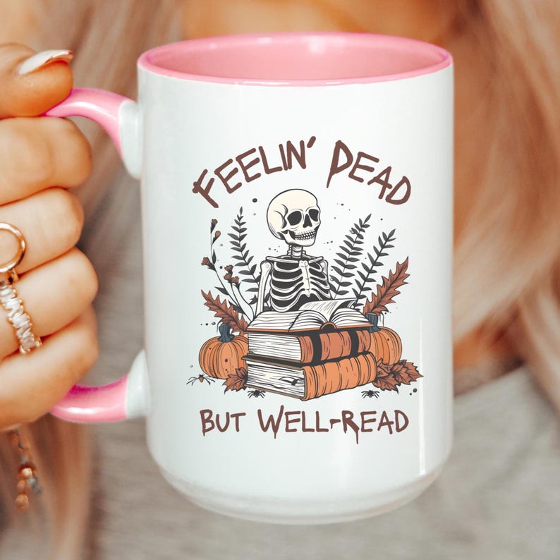 A white mug with a black or pink handle featuring a skeleton reading a book, surrounded by fall leaves and pumpkins, with the text: Feelin' Dead But Well-Read. Main colors are brown and orange.