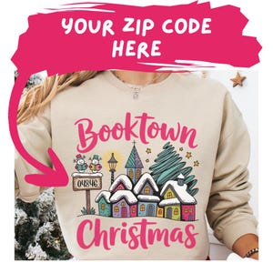 Puede incluir: Sudadera beige con un gráfico colorido que dice "Booktown Christmas". El gráfico presenta una escena de pueblo invernal con casas, una iglesia, un árbol de Navidad y muñecos de nieve. El texto "Your Zip Code Here" está encima del gráfico.
