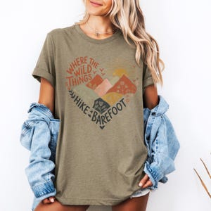 T-shirt with a heart-shaped mountain and floral design, sun rising in the background. Text reads: Where the Wild Things Hike Barefoot. The main colors are orange, green, red and yellow tones.