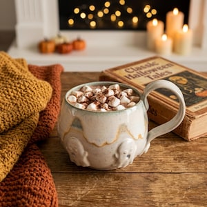 Può includere: Una tazza in ceramica con smalto bianco sporco maculato e figure di fantasmi in rilievo, riempita di cioccolata calda con mini marshmallow. La tazza presenta un manico curvo ed è posizionata accanto a un libro intitolato Classic Halloween Tales.