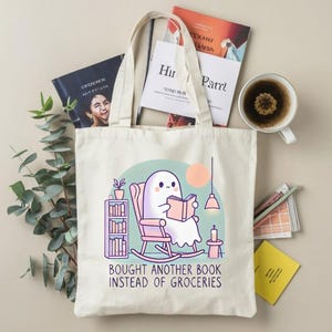 A natural-colored tote bag featuring a cute ghost reading a book with a humorous message about choosing books over groceries. The text says: Bought Another Book Instead Of Groceries. Main colors are blue, orange, white, pink and black.