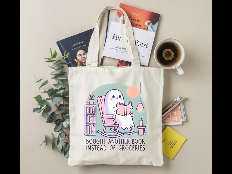 A natural-colored tote bag featuring a cute ghost reading a book with a humorous message about choosing books over groceries. The text says: Bought Another Book Instead Of Groceries. Main colors are blue, orange, white, pink and black.