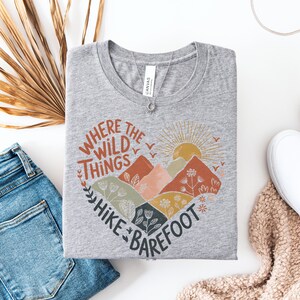 T-shirt with a heart-shaped mountain and floral design, sun rising in the background. Text reads: Where the Wild Things Hike Barefoot. The main colors are orange, green, red and yellow tones.