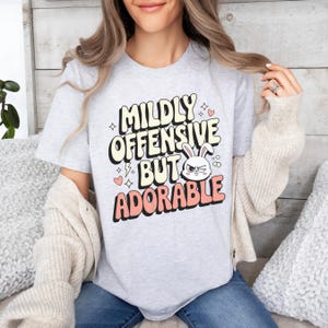 Funny graphic T-shirt featuring a sassy bunny with the phrase "Mildly Offensive But Adorable," perfect for humor-loving animal lovers. The main colors are white, beige and peach.