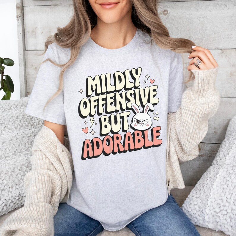 Funny graphic T-shirt featuring a sassy bunny with the phrase "Mildly Offensive But Adorable," perfect for humor-loving animal lovers. The main colors are white, beige and peach.