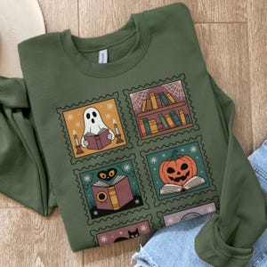 Może przedstawiać: Zielona bluza z czterema ilustracjami w stylu znaczków pocztowych o tematyce Halloween. Znaczki przedstawiają czytającego ducha, półkę z książkami, dynię i czarnego kota, wszystkie z motywem książki.