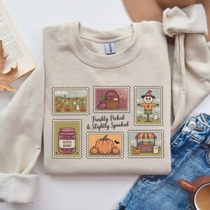 Op de afbeelding: Beige crewneck sweatshirt met een kleurrijke afbeelding van Halloween-thema postzegels. De postzegels tonen pompoenen, geesten, een vogelverschrikker en de tekst "Freshly Picked & Slightly Spooked".