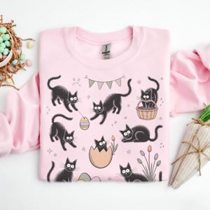 Puede incluir: Una sudadera rosa claro con un patrón repetido de gatos negros de dibujos animados. Los gatos se muestran en varias poses, algunos con huevos de Pascua y una cesta. El diseño incluye colores pastel y una pequeña pancarta.