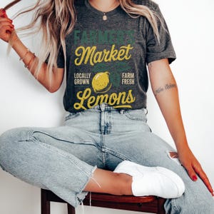 T-shirt with vintage-style “Farmer’s Market Lemons” graphic featuring a yellow lemon with leaves and rustic text. The main colors are yellow and green.