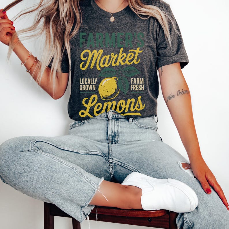 T-shirt with vintage-style “Farmer’s Market Lemons” graphic featuring a yellow lemon with leaves and rustic text. The main colors are yellow and green.