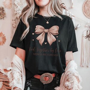 A sentimental t-shirt featuring a beautifully illustrated silk bow with the phrase: Tied Together by Love – Mom & Me Forever. Small hearts surround the design, making it a sweet and emotional tribute to the mother-daughter bond. The colors are very soft and all in pink tones.