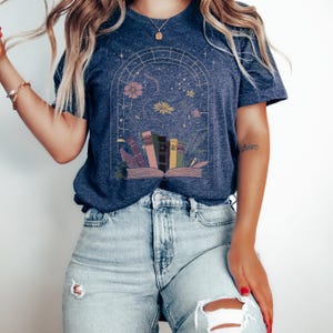 T-shirt featuring books, flowers, a snake, and stars framed by a mystical arched window—celestial, witchy, and dark academia styled.