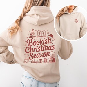 May include: A beige hoodie with a red graphic that says "Bookish Christmas Season." The design includes books, snowflakes, a Santa hat, and gifts. The hoodie has a front pocket and a hood.