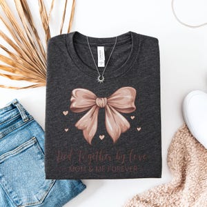 A sentimental t-shirt featuring a beautifully illustrated silk bow with the phrase: Tied Together by Love – Mom & Me Forever. Small hearts surround the design, making it a sweet and emotional tribute to the mother-daughter bond. The colors are very soft and all in pink tones.