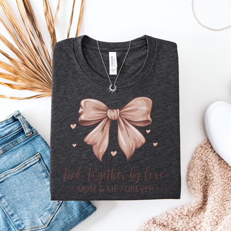 A sentimental t-shirt featuring a beautifully illustrated silk bow with the phrase: Tied Together by Love – Mom & Me Forever. Small hearts surround the design, making it a sweet and emotional tribute to the mother-daughter bond. The colors are very soft and all in pink tones.