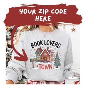 Op de afbeelding: Een lichtgrijs sweatshirt met de tekst "BOOK LOVERS TOWN" en een afbeelding van drie kleurrijke huizen en een kerstboom. Het sweatshirt heeft ook de tekst "YOUR ZIP CODE HERE" in een rode banner.