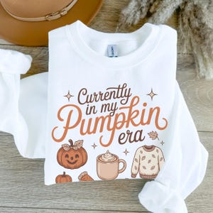 May include: White sweatshirt with the text "Currently in my Pumpkin Era" in brown and orange. The design includes a smiling pumpkin, a mug of coffee, and a sweater with leaves.