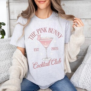 A trendy cocktail-themed t-shirt featuring a pink martini glass with a bunny accent and vintage lettering, perfect for bar lovers and aesthetic outfits. The main colors are pink and white.
