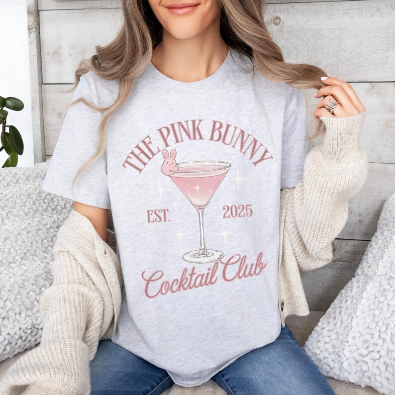 A trendy cocktail-themed t-shirt featuring a pink martini glass with a bunny accent and vintage lettering, perfect for bar lovers and aesthetic outfits. The main colors are pink and white.