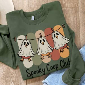 May include: An olive green sweatshirt featuring a graphic of three cartoon ghosts hanging on a washing line. The ghosts have bird-like bodies and are set against a backdrop of vertical stripes in muted colours. The text "Spooky Coop Club" is printed below.