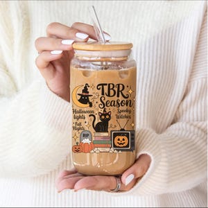 May include: A glass tumbler with a bamboo lid and straw, filled with a beverage. The tumbler features a Halloween-themed design with a black cat, pumpkins, and the text "TBR Season". The design is in orange, black, and white.