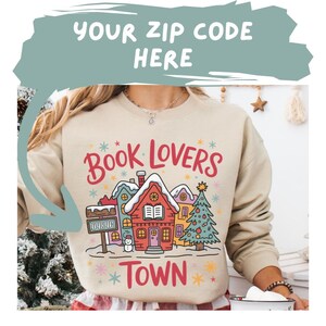 Puede incluir: Sudadera beige con un gráfico colorido de una escena de pueblo. El diseño incluye el texto "Book Lovers Town" en rojo y una fila de casas con un árbol de Navidad. El texto "Your Zip Code Here" está en la parte superior.