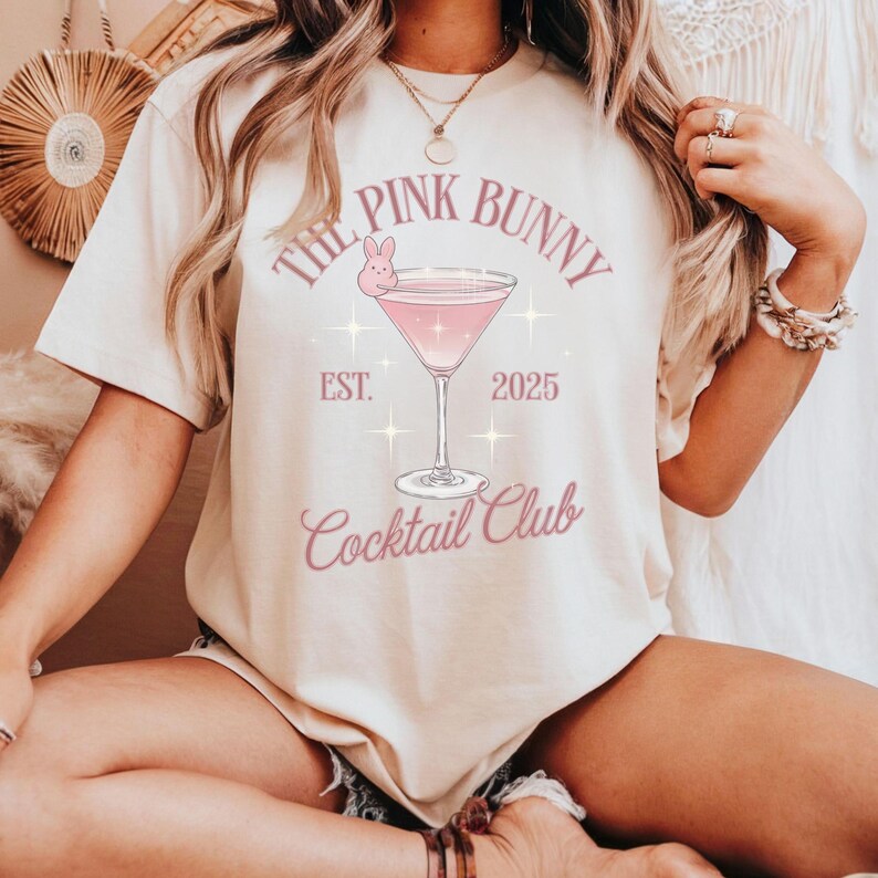 A trendy cocktail-themed t-shirt featuring a pink martini glass with a bunny accent and vintage lettering, perfect for bar lovers and aesthetic outfits. The main colors are pink and white.