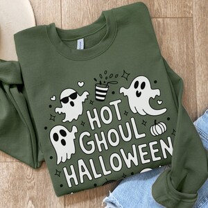 Può includere: Felpa verde oliva con grafica bianca "HOT GHOUL HALLOWEEN". Il design include fantasmi dei cartoni animati, una zucca e una bevanda con cannuccia. La felpa è piegata su una superficie di legno.