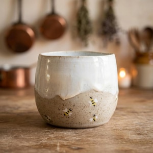 Op de afbeelding: Een keramische mok met een tweekleurig ontwerp, bestaande uit een glad wit geglazuurd bovendeel en een gespikkeld beige onderste deel. Het onderste gedeelte is versierd met kleine, handgeschilderde gele en zwarte bijen op het oppervlak.