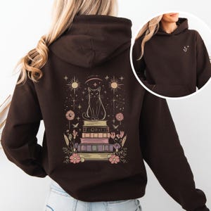Puede incluir: Sudadera con capucha marrón oscuro con un diseño de fantasía en la espalda. El diseño muestra un gato sobre libros, con estrellas, soles y flores. Un diseño más pequeño está en la parte delantera, cerca de la capucha.