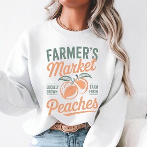 May include: White sweatshirt with a graphic design that says "Farmer's Market Peaches" with two peaches and green leaves. The text "Locally Grown" and "Farm Fresh" are on either side of the peaches.