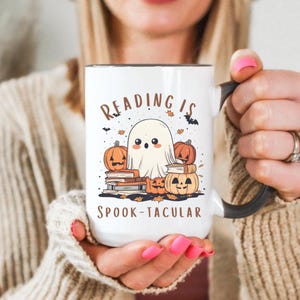 Op de afbeelding: Witte keramische mok met een zwart handvat en rand, met een Halloween-thema ontwerp. Het ontwerp bevat een cartoon spook, pompoenen, boeken en de tekst "READING IS SPOOK-TACULAR" in bruine letters. De mok wordt door een persoon vastgehouden.