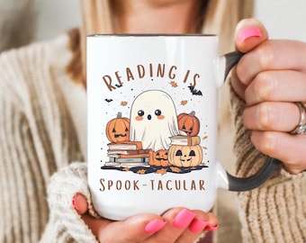 Può includere: Tazza in ceramica bianca con manico e bordo neri, con un design a tema Halloween. Il design include un fantasma dei cartoni animati, zucche, libri e il testo "READING IS SPOOK-TACULAR" in lettere marroni. La tazza è tenuta da una persona.