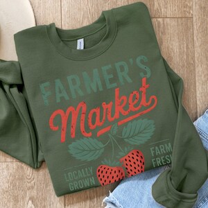 May include: Green sweatshirt with the text "FARMER'S Market" in red and green lettering. The text is distressed and has a vintage look. There are two red strawberries with green leaves on the bottom of the sweatshirt. The text "LOCALLY GROWN" and "FARM FRESH" are also on the sweatshirt.